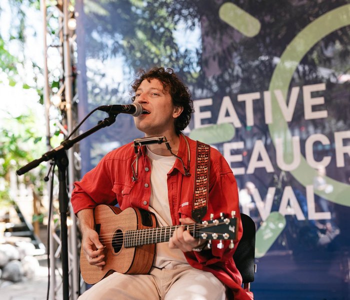 Eine Person spielt Gitarre und singt mit einem Lächeln auf der Bühne. Im Hintergrund ist ein CBF-Banner zu sehen.
