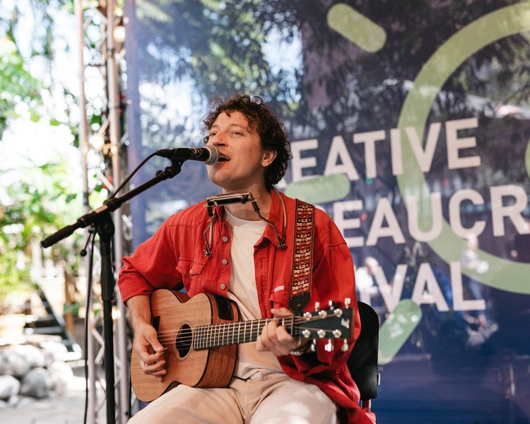 Eine Person spielt Gitarre und singt mit einem Lächeln auf der Bühne. Im Hintergrund ist ein CBF-Banner zu sehen.