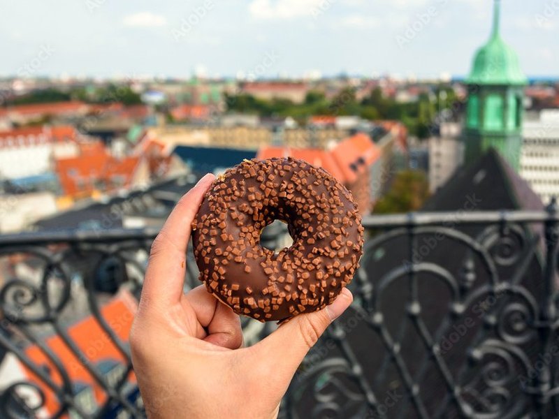 Eine Hand hält einen Donut, im Hintergrund ein Blick von einer Terrasse über die Dächer der Stadt.