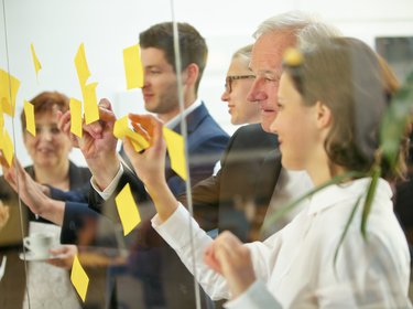 Kolleginnen und Kollegen stehen vor einer Glaswand und kleben gelbe Notizzettel