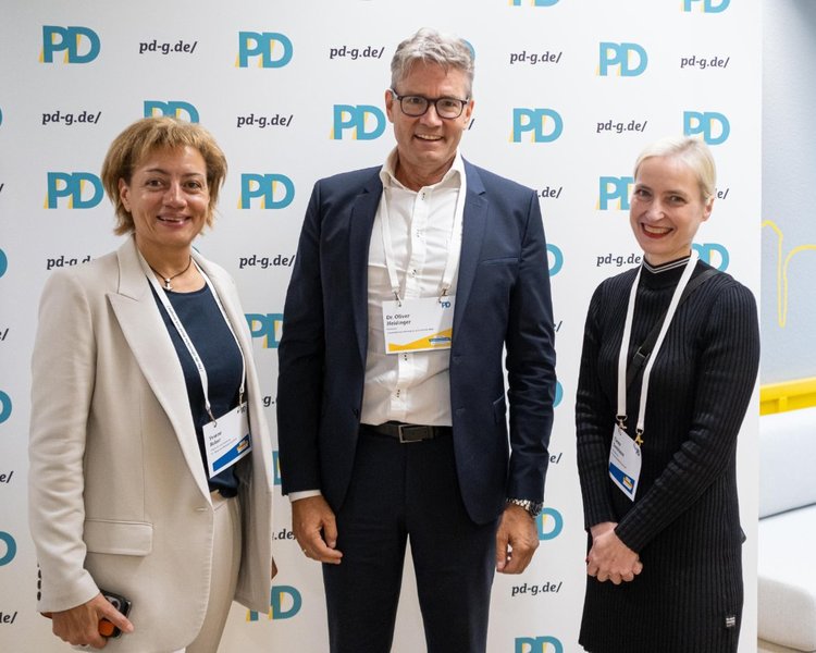Yvonne Balzer (PD-Standortleitung), Dr. Oliver Heidinger (Präsident, Landesbetrieb Information und Technik NRW) und Theresa Twachtmann (Geschäftsführerin der PD) vor einer PD-Logo-Wand