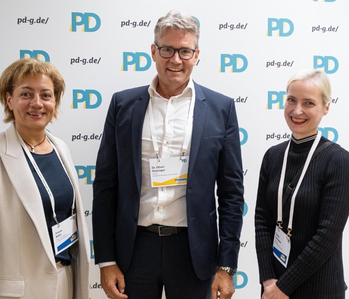 Yvonne Balzer (PD-Standortleitung), Dr. Oliver Heidinger (Präsident, Landesbetrieb Information und Technik NRW) und Theresa Twachtmann (Geschäftsführerin der PD) vor einer PD-Logo-Wand