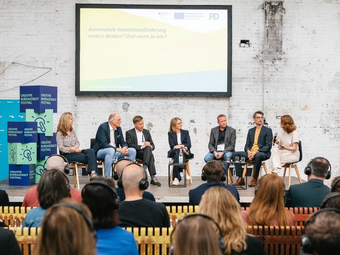 Eine Panel-Diskussion auf einer großen Bühne. Das Publikum ist ihnen zugewandt und einige tragen Kopfhörer für die Sprachübersetzung. Im Hintergrund ist eine Präsentation zu sehen.
