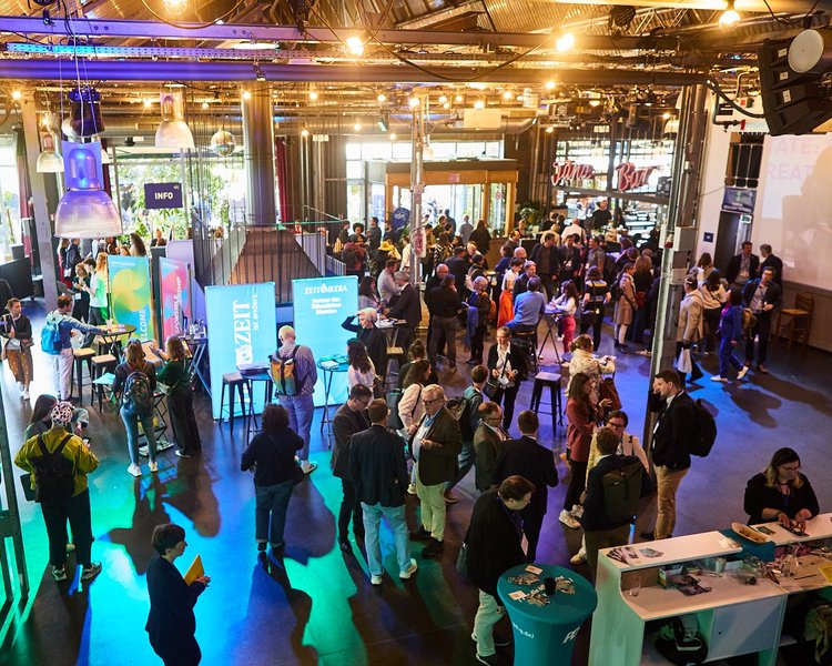 Ein Saal voller Festivalbesuchenden, die sich an den Ständen der Partner des CBFs austauschen. Bunte Lichter tauchen den Raum in eine lebendige Atmosphäre.
