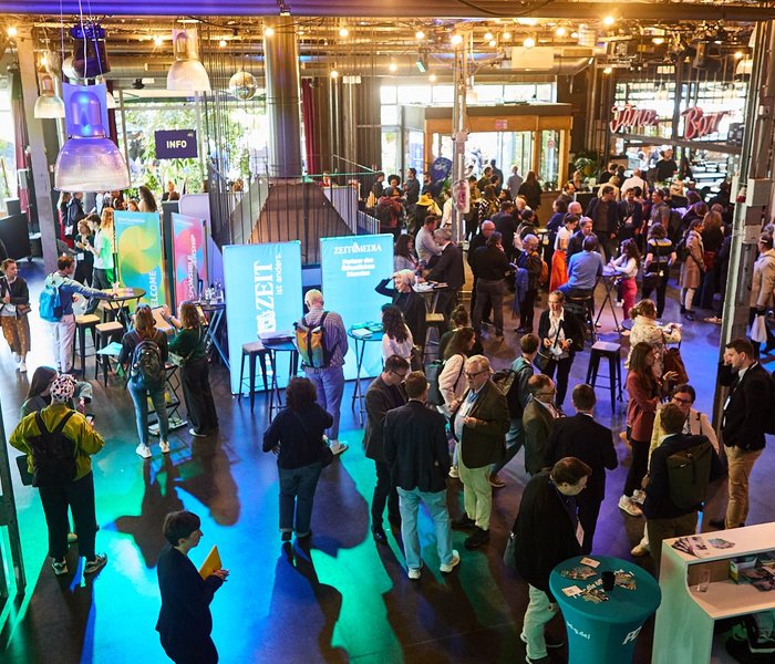 Ein Saal voller Festivalbesuchenden, die sich an den Ständen der Partner des CBFs austauschen. Bunte Lichter tauchen den Raum in eine lebendige Atmosphäre.