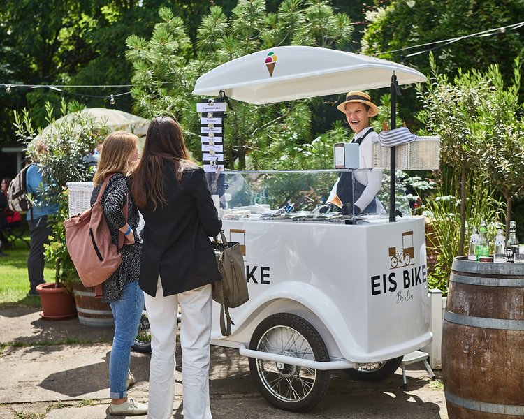 Eine lachende Eisverkäuferin bedient zwei Personen an einem mobilen Eiswagenstand. Sie sind draußen im Grünen.