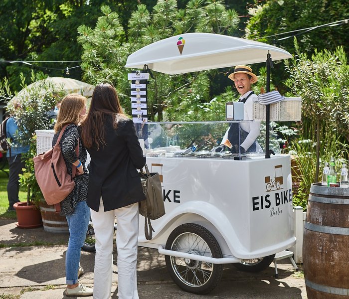 Eine lachende Eisverkäuferin bedient zwei Personen an einem mobilen Eiswagenstand. Sie sind draußen im Grünen.