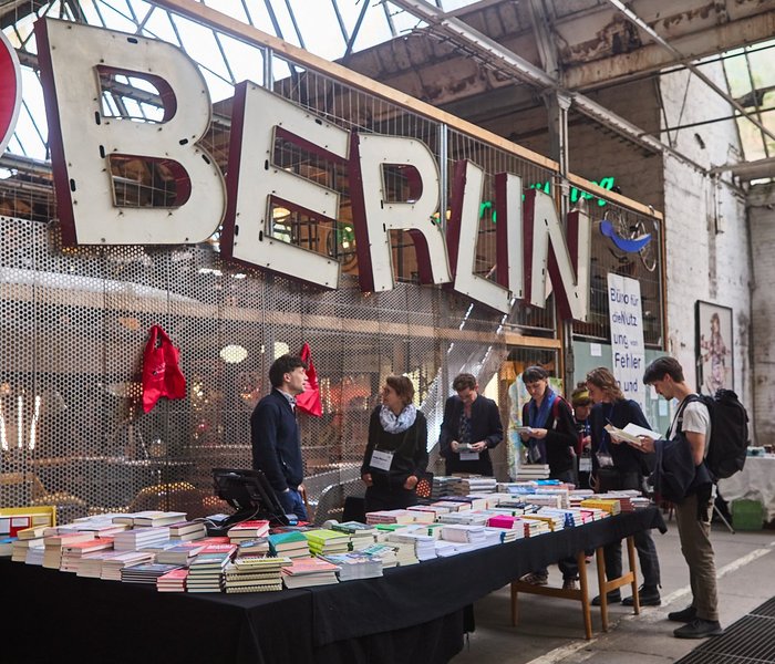 Ein großer Buchstand auf dem CBF, um den Personen stehen. Einige lesen in Büchern, während die Person hinter dem Stand sich mit einer weiteren Person unterhält.