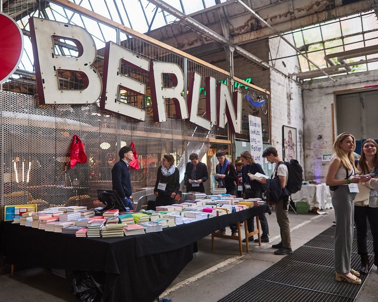 Ein großer Buchstand auf dem CBF, um den Personen stehen. Einige lesen in Büchern, während die Person hinter dem Stand sich mit einer weiteren Person unterhält.