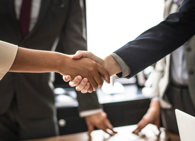 Zwei Personen geben sich die Hand, zwei weitere im Hintergrund, Besprechungssituation