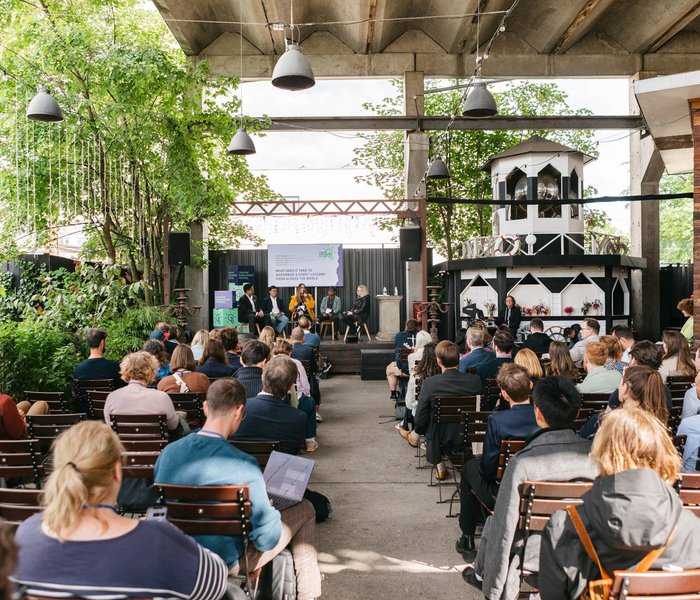 Menschen sitzen in Reihen vor einer Außenbühne, auf der eine Panel-Diskussion stattfindet. An den Seiten der Bühne sind Bäume und Bambus zu sehen.