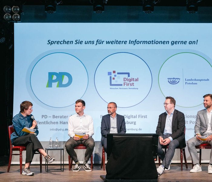 Auf einer Bühne sitzen Personen und halten eine Panel-Diskussion. Mit dabei sind PD - Beratung der öffentlichen Hand, Landeshauptstadt Potsdam und Hansestadt Hamburg mit dem Projekt "DigitalFirst".