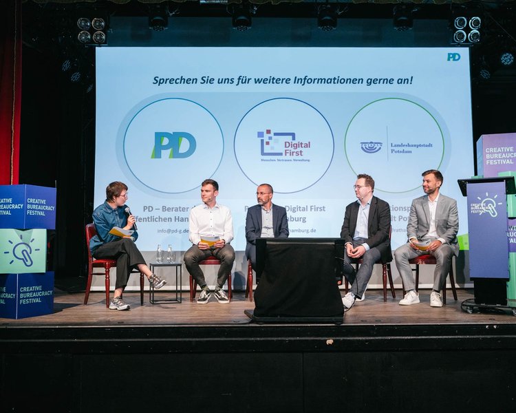Auf einer Bühne sitzen Personen und halten eine Panel-Diskussion. Mit dabei sind PD - Beratung der öffentlichen Hand, Landeshauptstadt Potsdam und Hansestadt Hamburg mit dem Projekt "DigitalFirst".